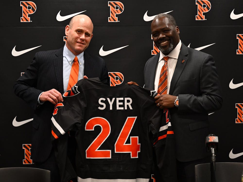 Two men posing with a hockey jersey at a press conference with the Nike and Princeton logos displayed in the background 