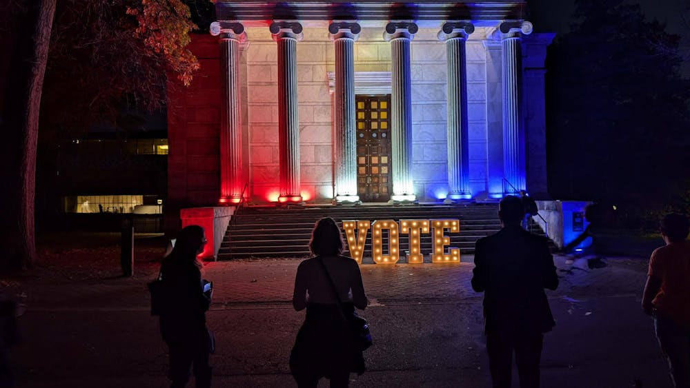 A white colonial-style building is lit up with blue and red lights, and a large lit-up "VOTE" sign is propped up in the front. Multiple people gather in the foreground.