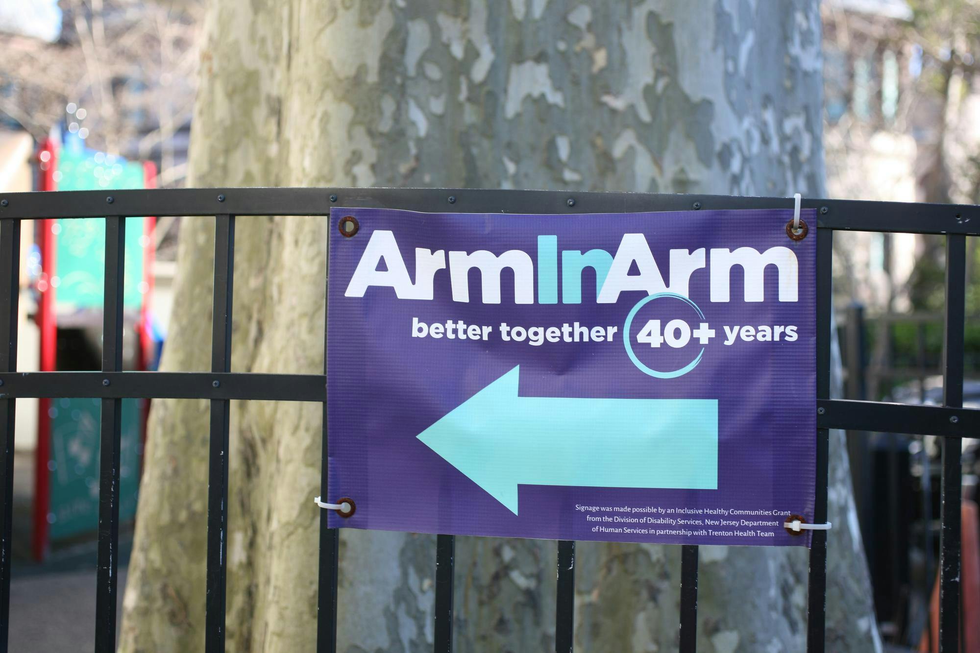 An indigo canvas sign zip-tied to a black fence reads "Arm In Arm, better together, 40+ years" in white and turquoise lettering. A turquoise arrow points to the left. A tree and playground are blurred in the background.