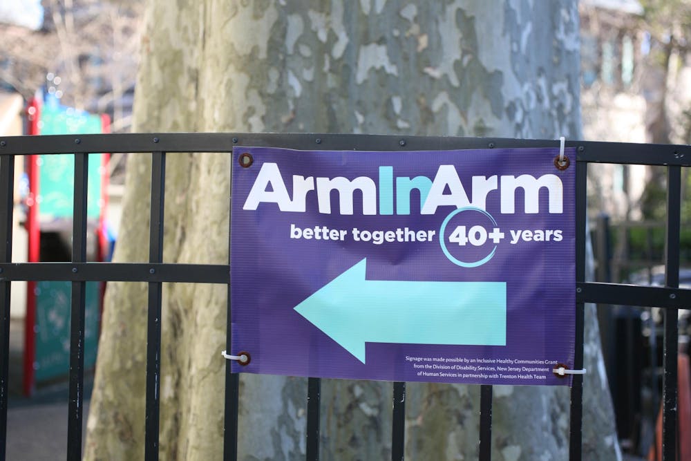 An indigo canvas sign zip-tied to a black fence reads "Arm In Arm, better together, 40+ years" in white and turquoise lettering. A turquoise arrow points to the left. A tree and playground are blurred in the background.