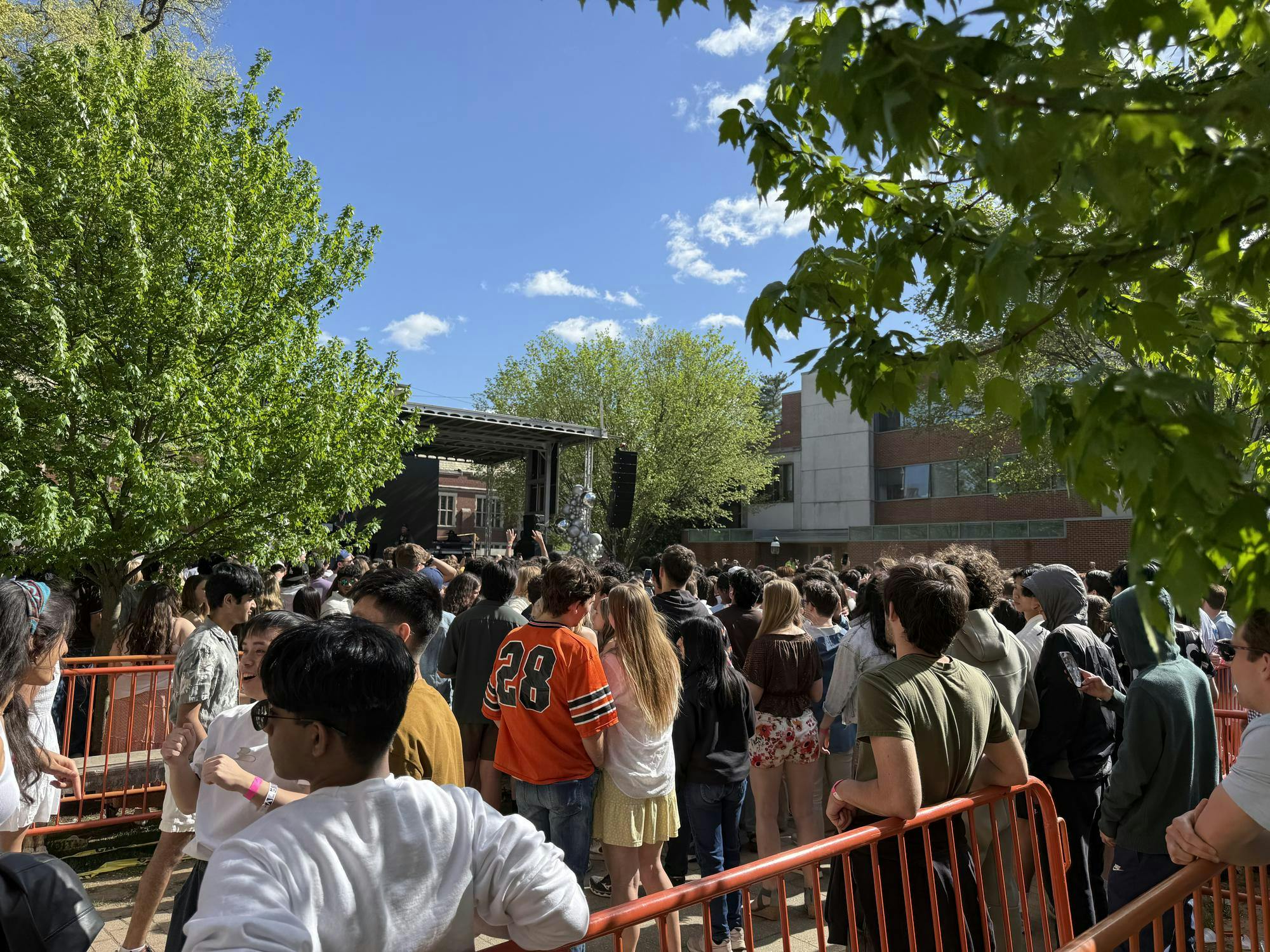 A large crowd of people gather on a lawn. There is an outdoor stage in the background with trees surrounding the lawn.