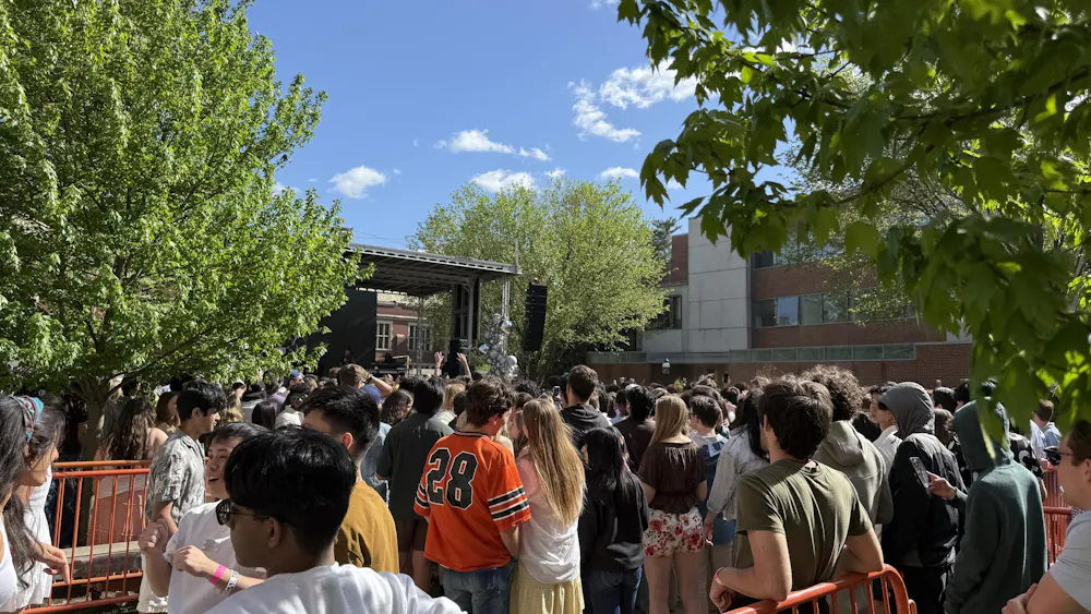 A large crowd of people gather on a lawn. There is an outdoor stage in the background with trees surrounding the lawn.