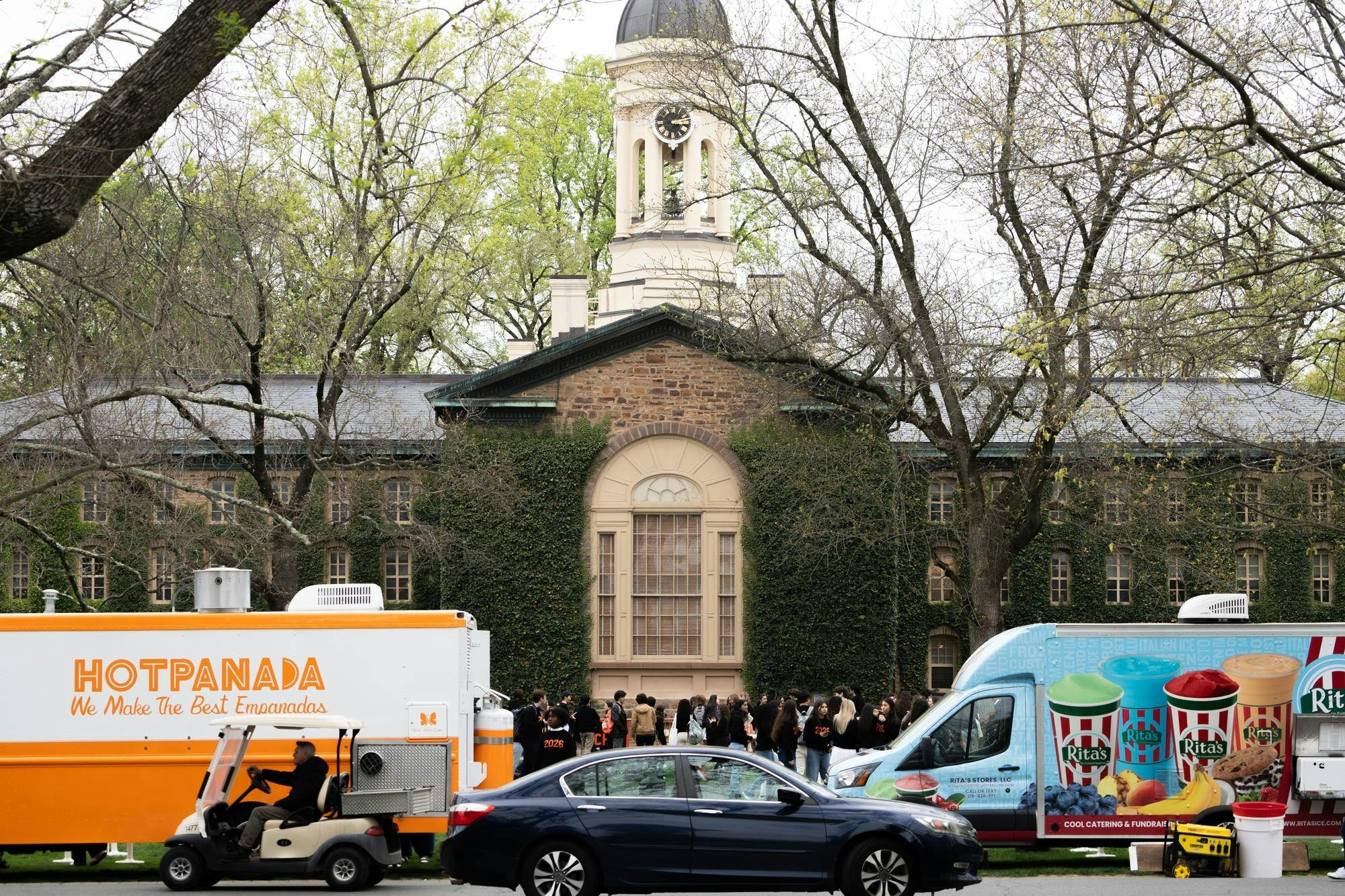 Two food trucks flank a crowd of students wearing black and orange sweaters in front of an ivy-covered building.