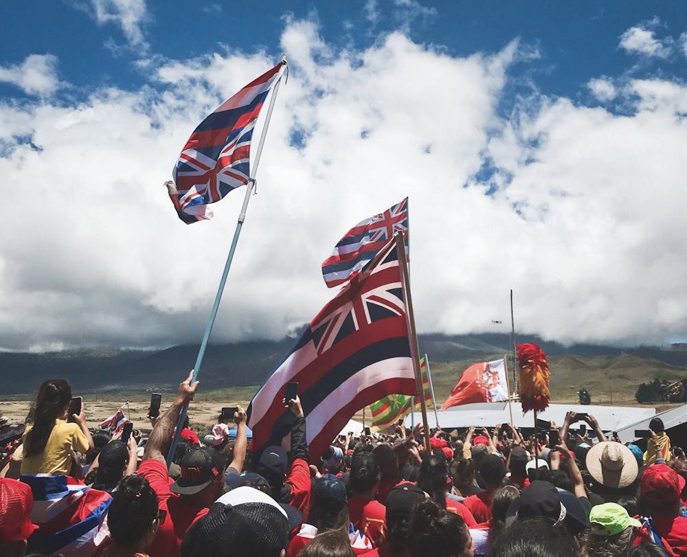 <h5>2019 – <em>Kia’i </em>(protectors) wave inverted Hawaiian flags to illustrate a nation in distress; their hands, which are in a “triangle shape” to indicate a shape of a mountain, highlight their support and love for the Hawaiian people’s sacred <a href="https://www.honolulumagazine.com/the-sacred-history-of-maunakea/"><u>Mauna Kea</u></a> during the Thirty-Meter Telescope (<a href="https://www.hawaiinewsnow.com/2019/07/13/exploring-timeline-leading-up-conflict-mauna-kea/"><u>TMT</u></a>) protests.&nbsp;</h5>
<h6>Photo Courtesy of Monet Bisch&nbsp;</h6>