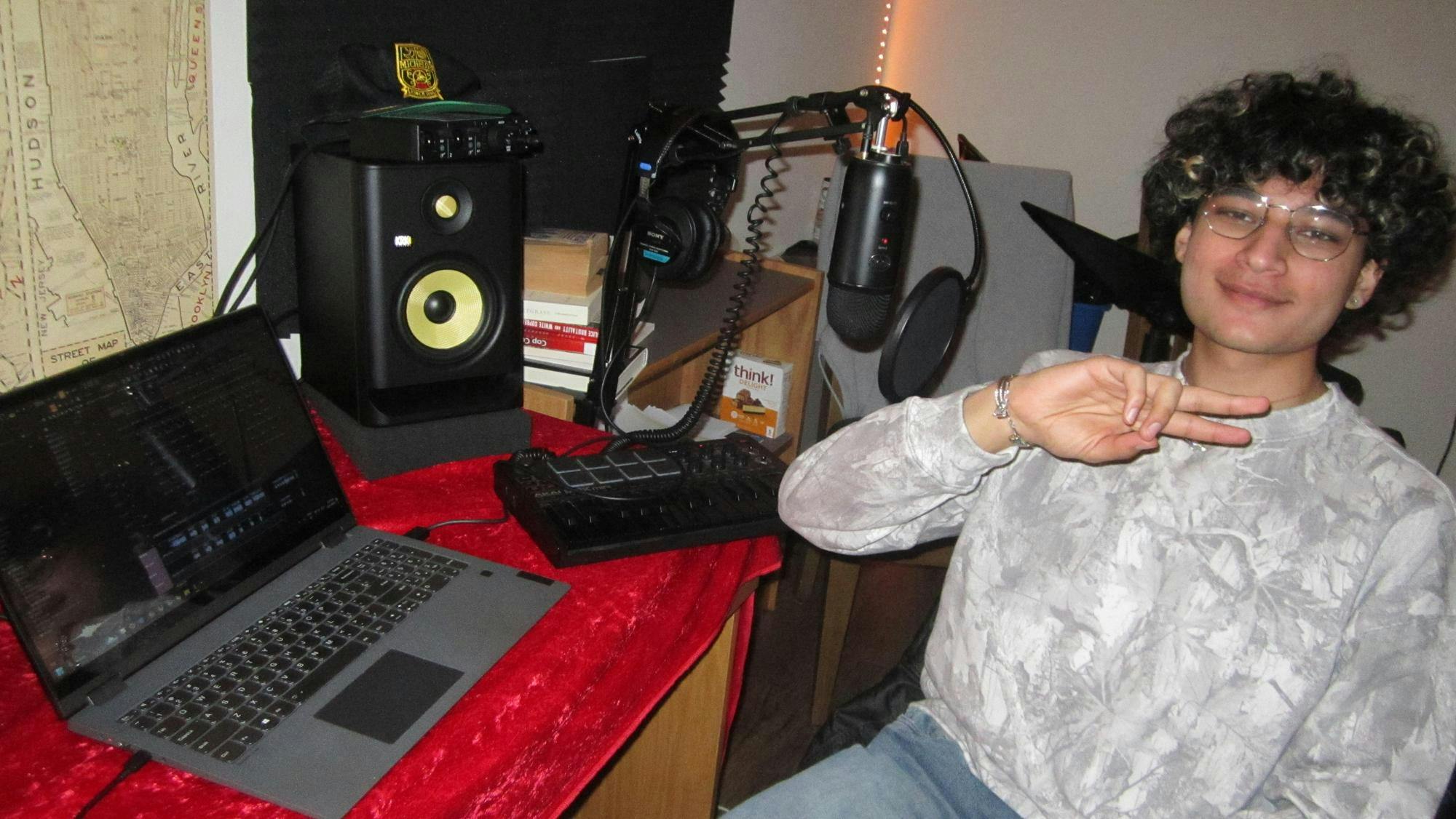 A boy with curly hair and a camouflage crewneck sits in-front of a laptop, speaker, and DJ board. 