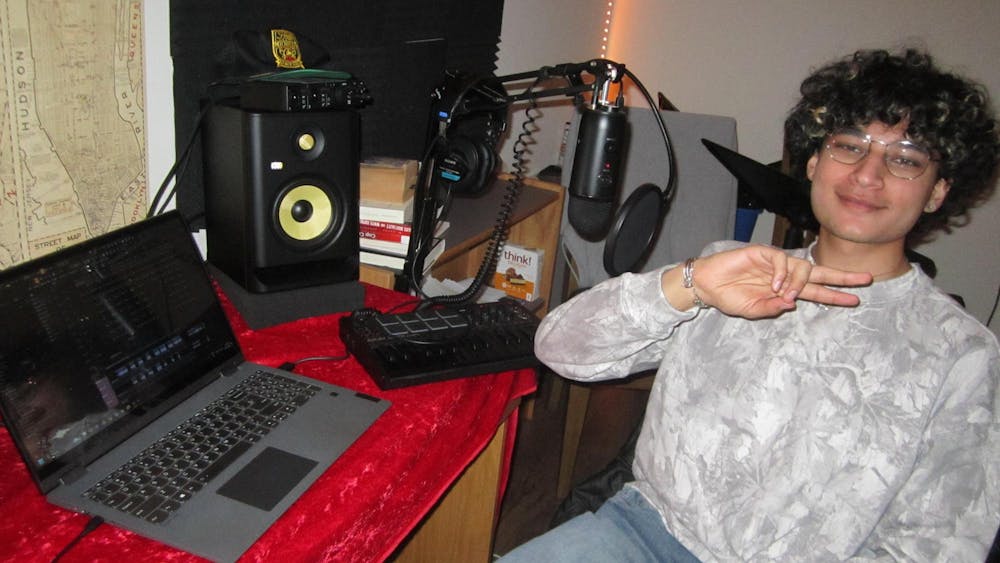 A boy with curly hair and a camouflage crewneck sits in-front of a laptop, speaker, and DJ board. 