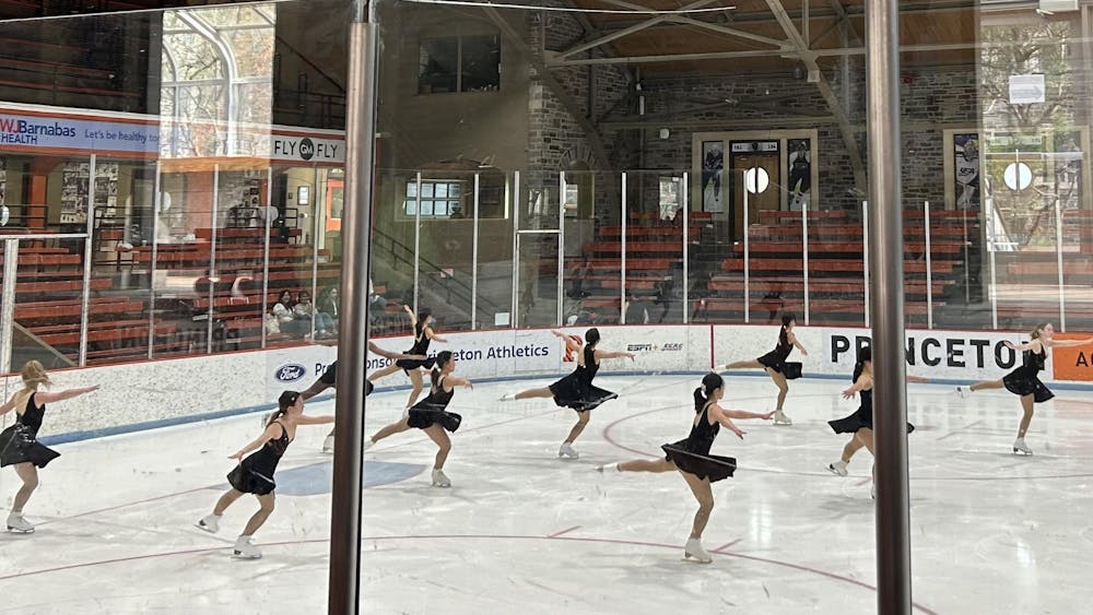A formation of ice skaters with black costumes twirl on the ice. 