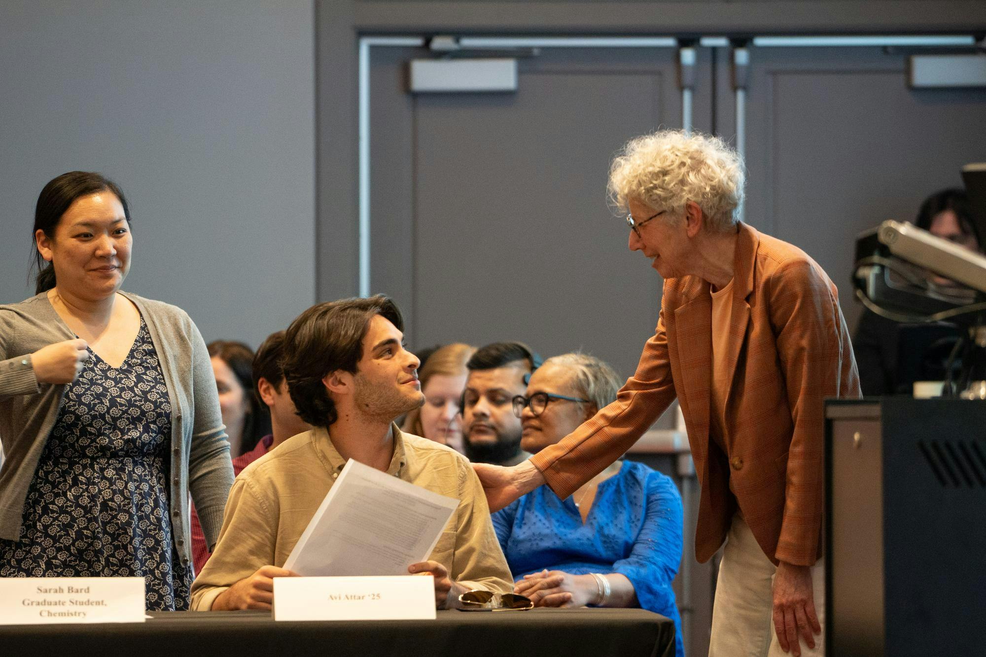 Two people speak in a gray room.