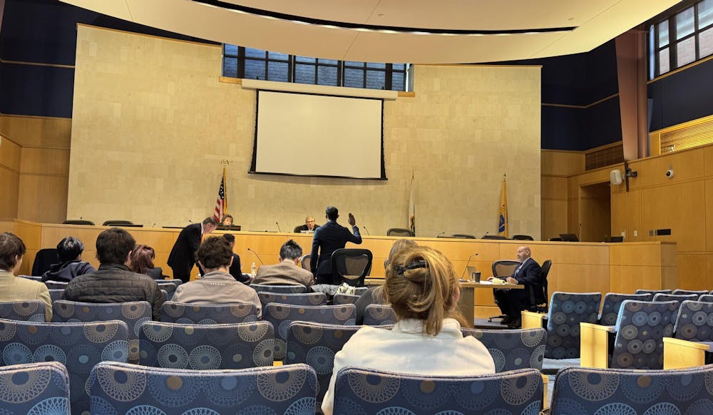 The photo is of a courtroom, taken from a seat facing the judge, defense, and prosecution. A man in a suit stands in the center with his hand raised, taking an oath to say the truth.