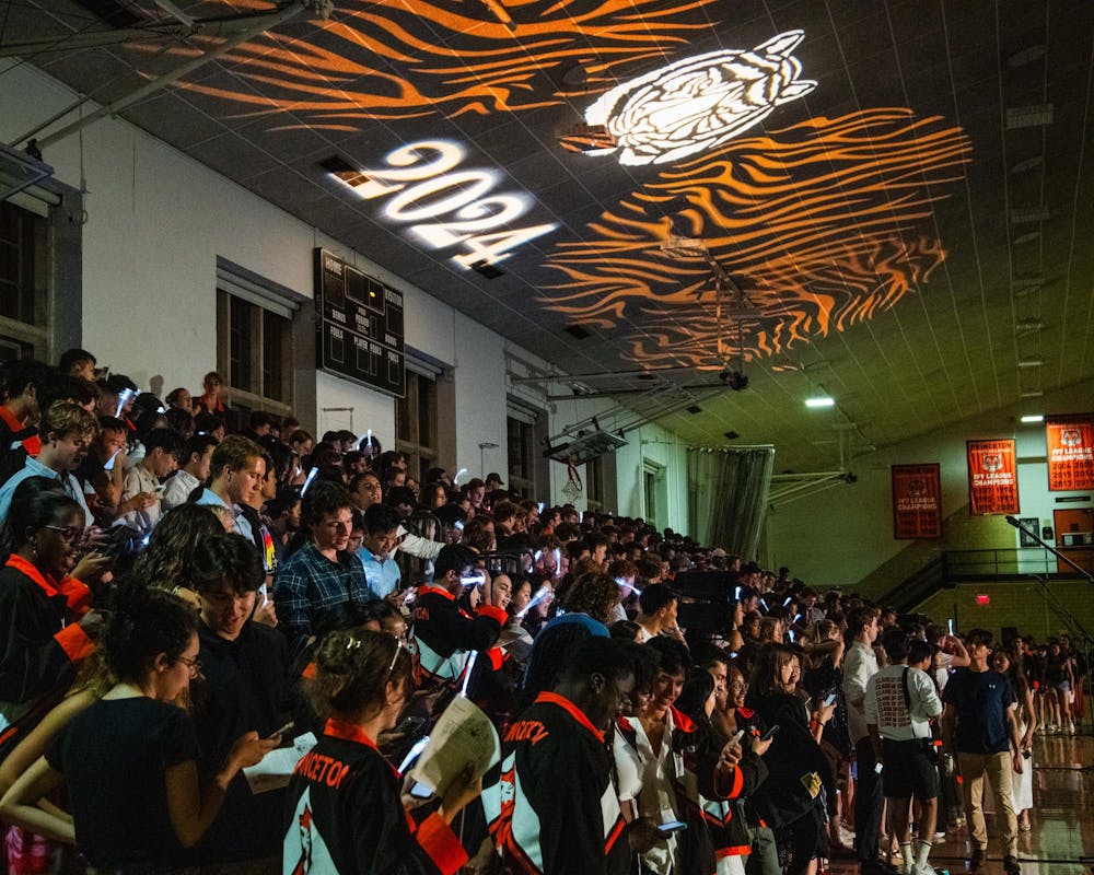A wide array of Princeton graduates, many of whom are wearing black and orange jackets, line bleachers inside a dim gymnasium.