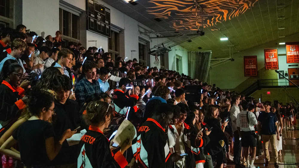 A wide array of Princeton graduates, many of whom are wearing black and orange jackets, line bleachers inside a dim gymnasium.