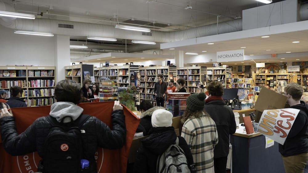 About fifteen people are shown gathered around a table of books, with one person holding a microphone.