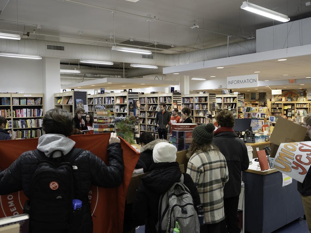 About fifteen people are shown gathered around a table of books, with one person holding a microphone.