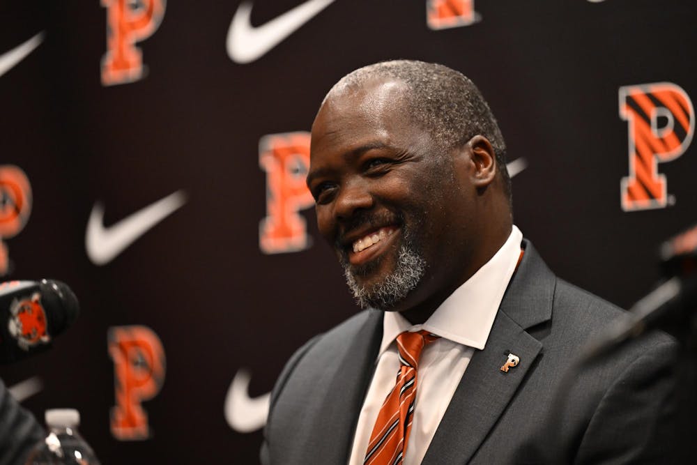 Man in a dark suit and orange tie, smiling, standing in front of a black media backdrop patterned with orange Princeton University logos and white Nike logos. 
