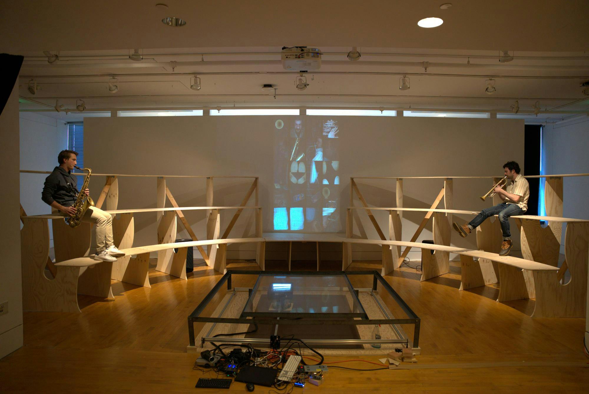The setup of the exhibition, with wooden benches surrounding the scanner in the center. There are scans projected onto a wall behind the benches. 
