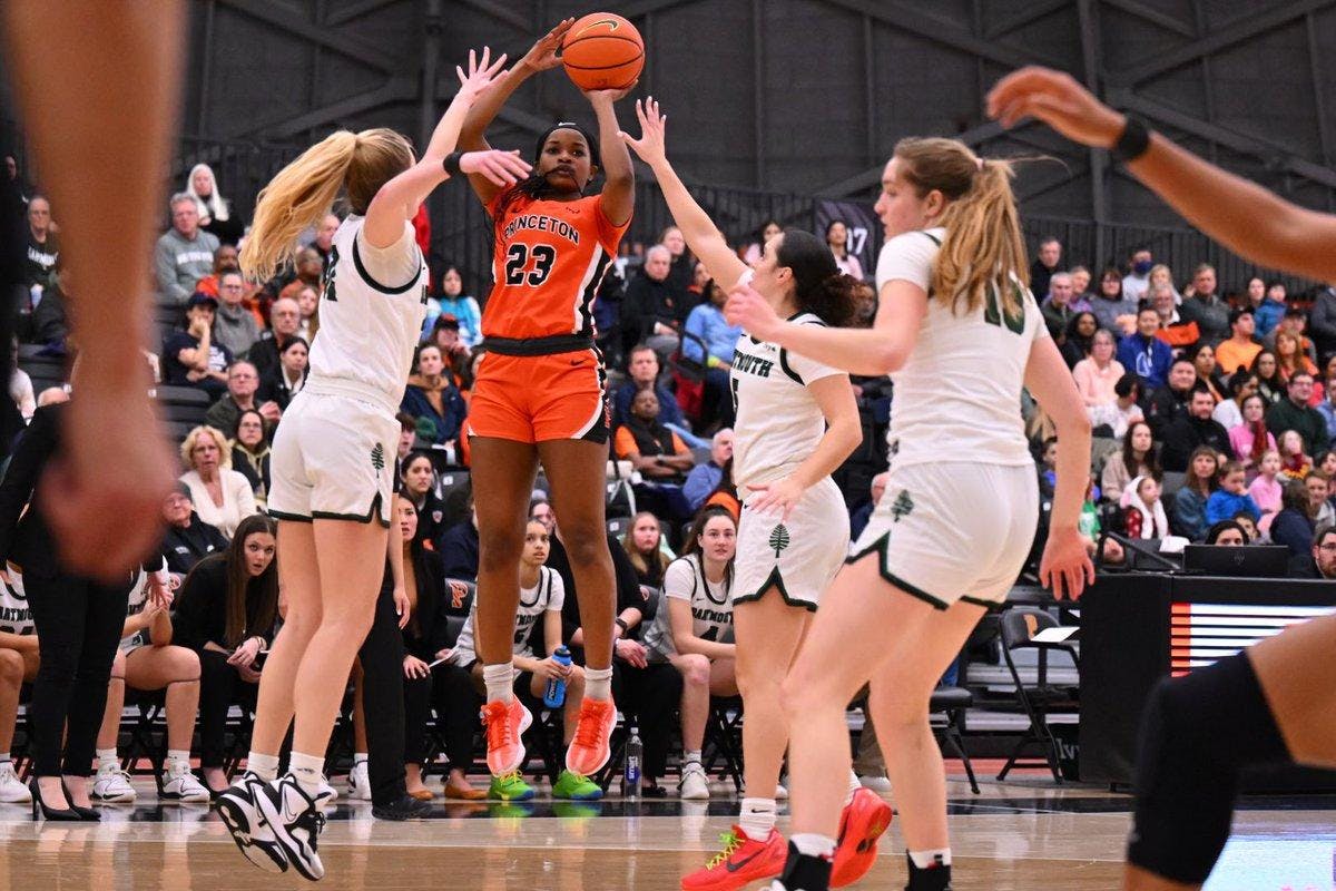 Player in orange and black Princeton jersey shoots a three pointer over two Dartmouth defenders. 
