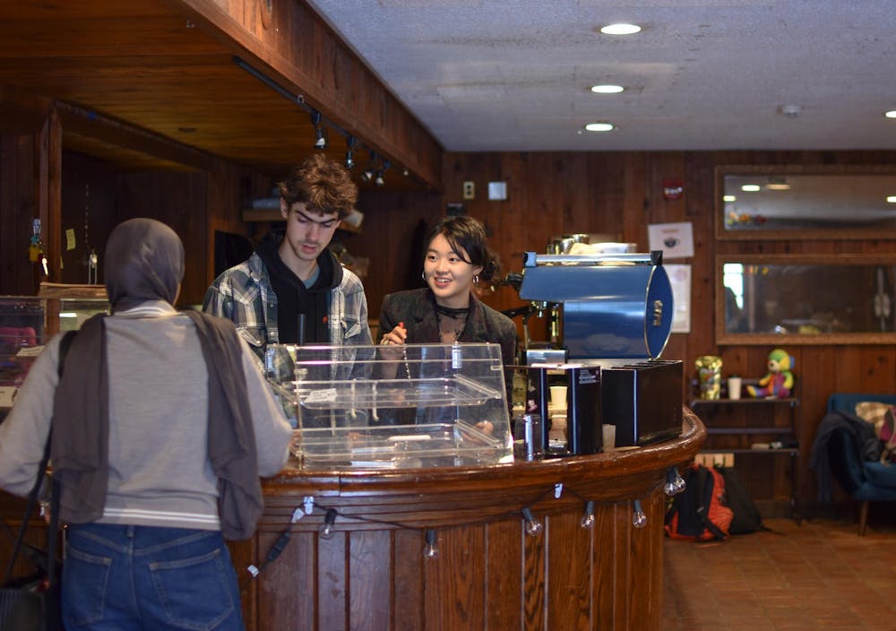 Two student baristas stand behind a wooden counter. Behind the wooden counter is a steel expresso machine. One student stands in front of the counter, interacting with the baristas behind the counter.