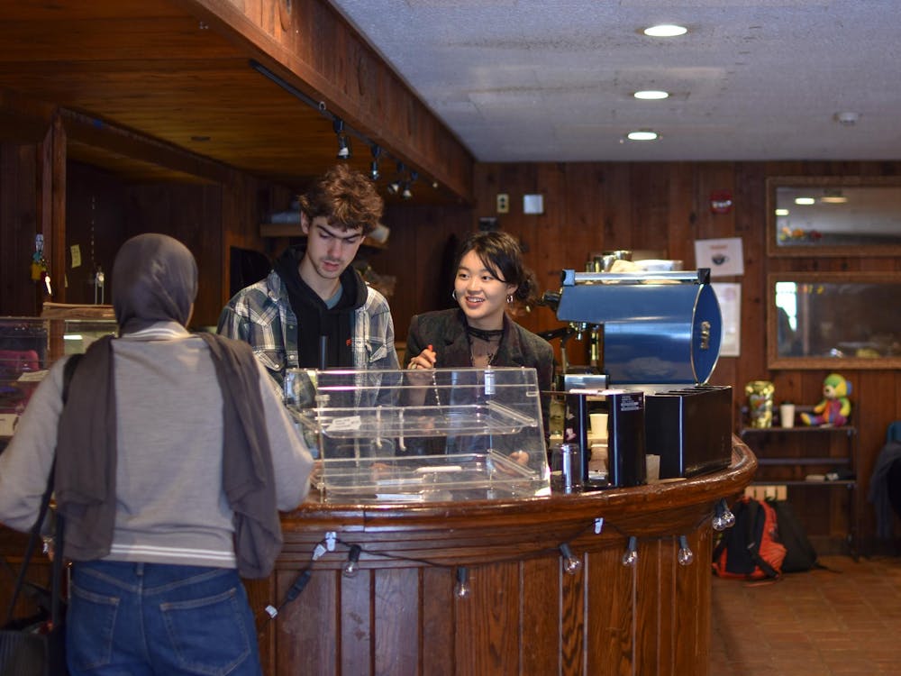 Two student baristas stand behind a wooden counter. Behind the wooden counter is a steel expresso machine. One student stands in front of the counter, interacting with the baristas behind the counter.