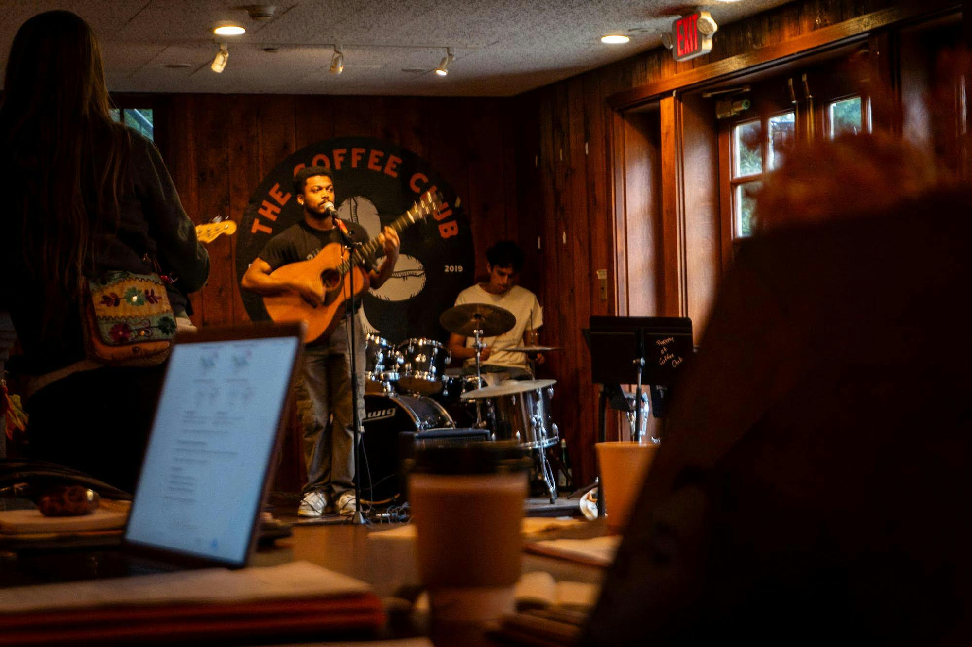A man in a black T-shirt plays guitar in a wooden room filled with amber light and many people. 