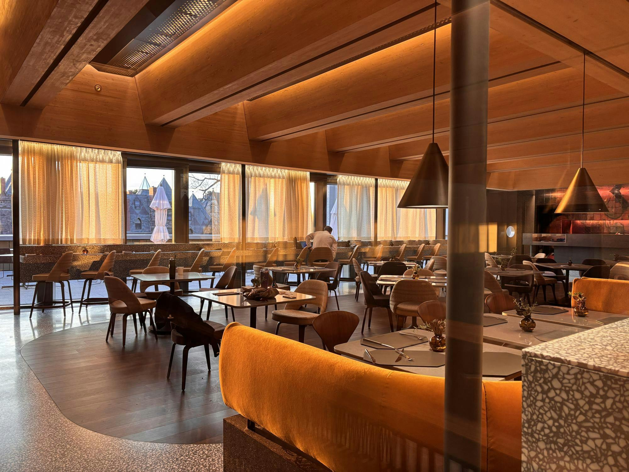 Behind a glass window, is a warmly lit orange room with yellow couches, brown tables, and chairs throughout. 