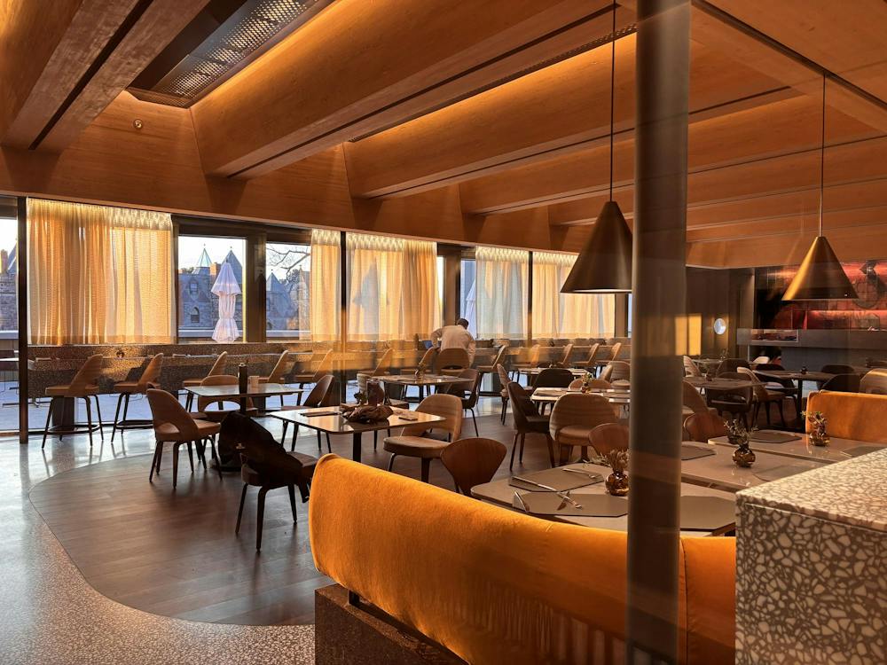 Behind a glass window, is a warmly lit orange room with yellow couches, brown tables, and chairs throughout. 