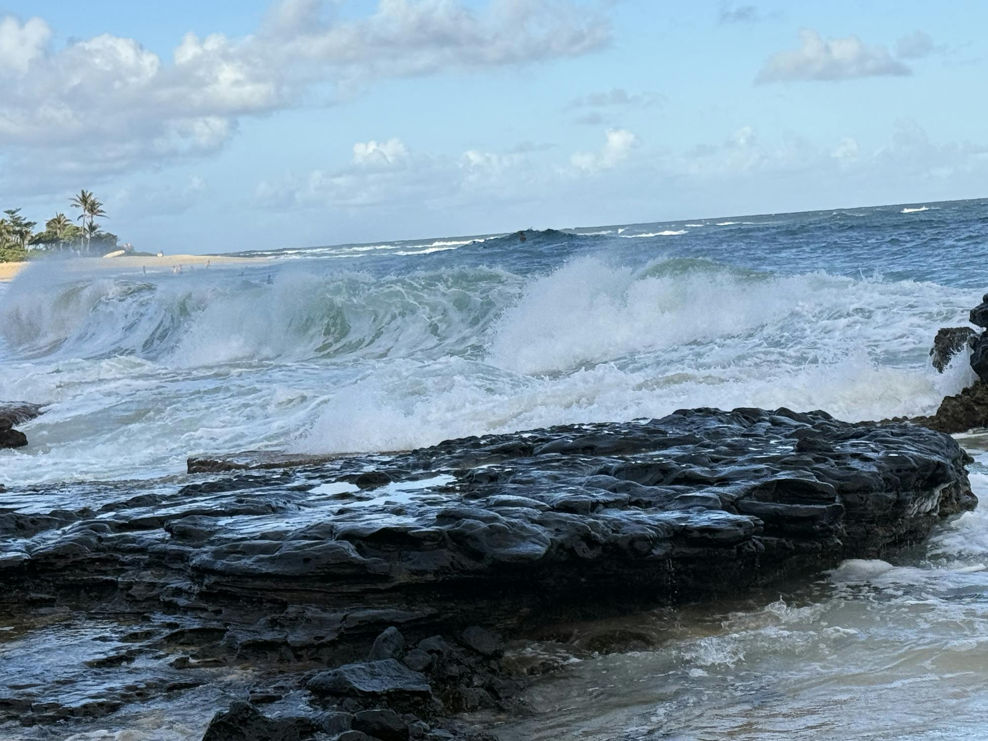 Foamy blue and white waves crash over black rock next to a sunny beach in background.