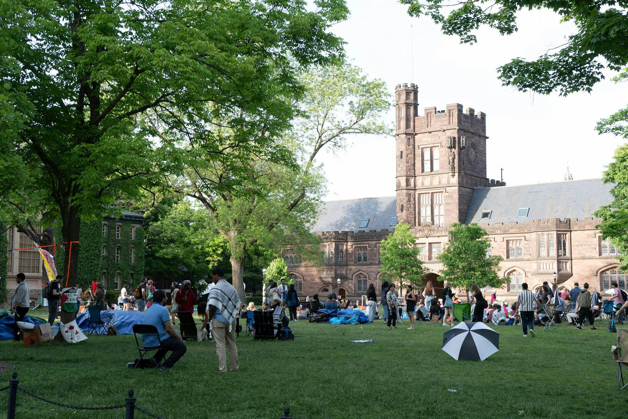 A group of people stand around a grassy courtyard. An old brick building stands in the background.