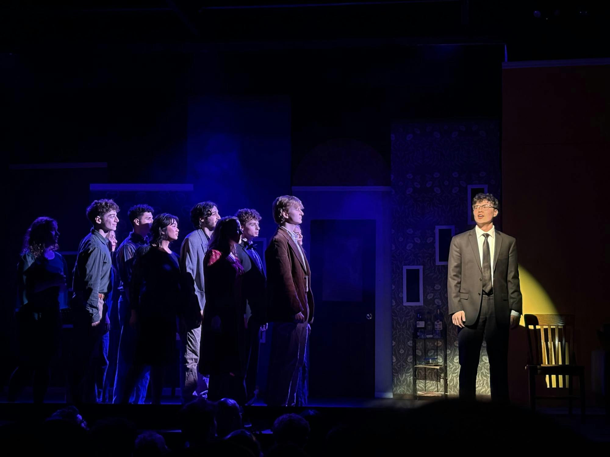 Actors stand on the left side of the stage, under a dark blue light, while one actor stands on the right side of the stage, under a spotlight. 