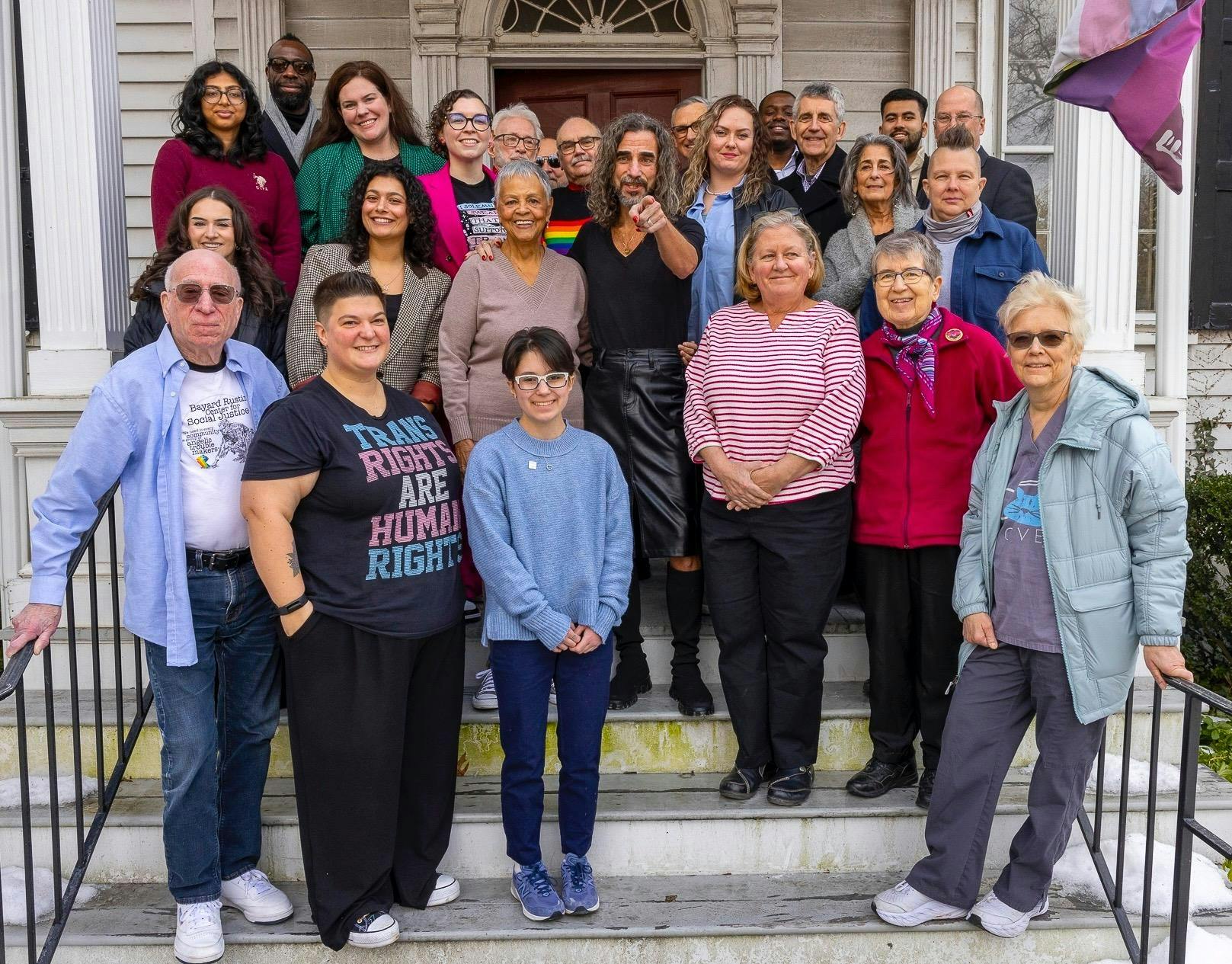 A group of around 20 people wearing colorful attire stand on the staircase of the house, smiling at the camera. 