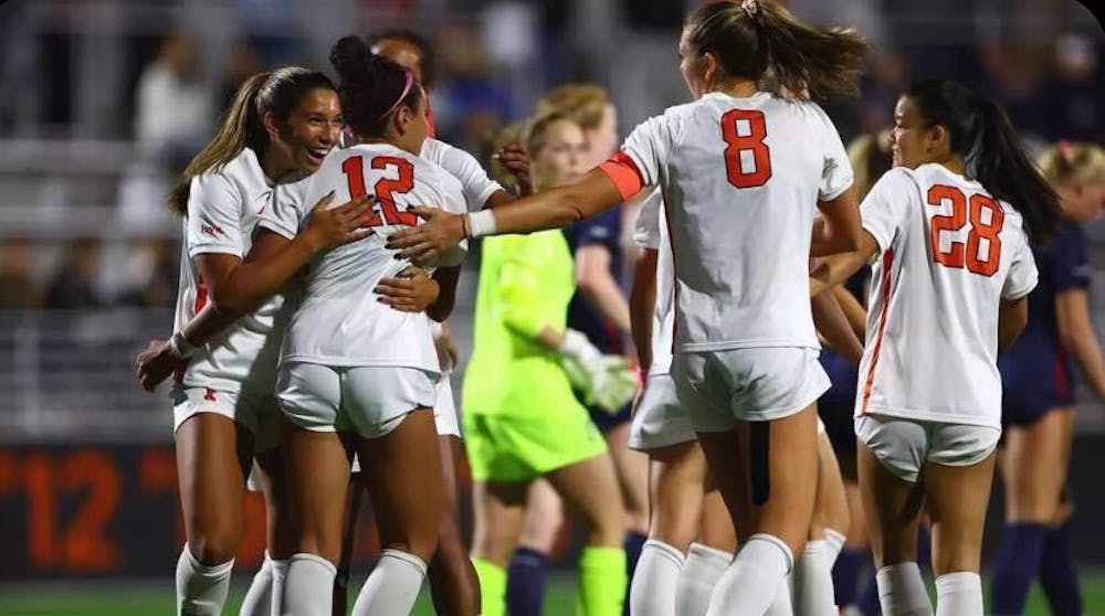 Four girls in white and orange white and orange soccer kits celebrate a goal. A goalkeeper in bright yellow stands in the background