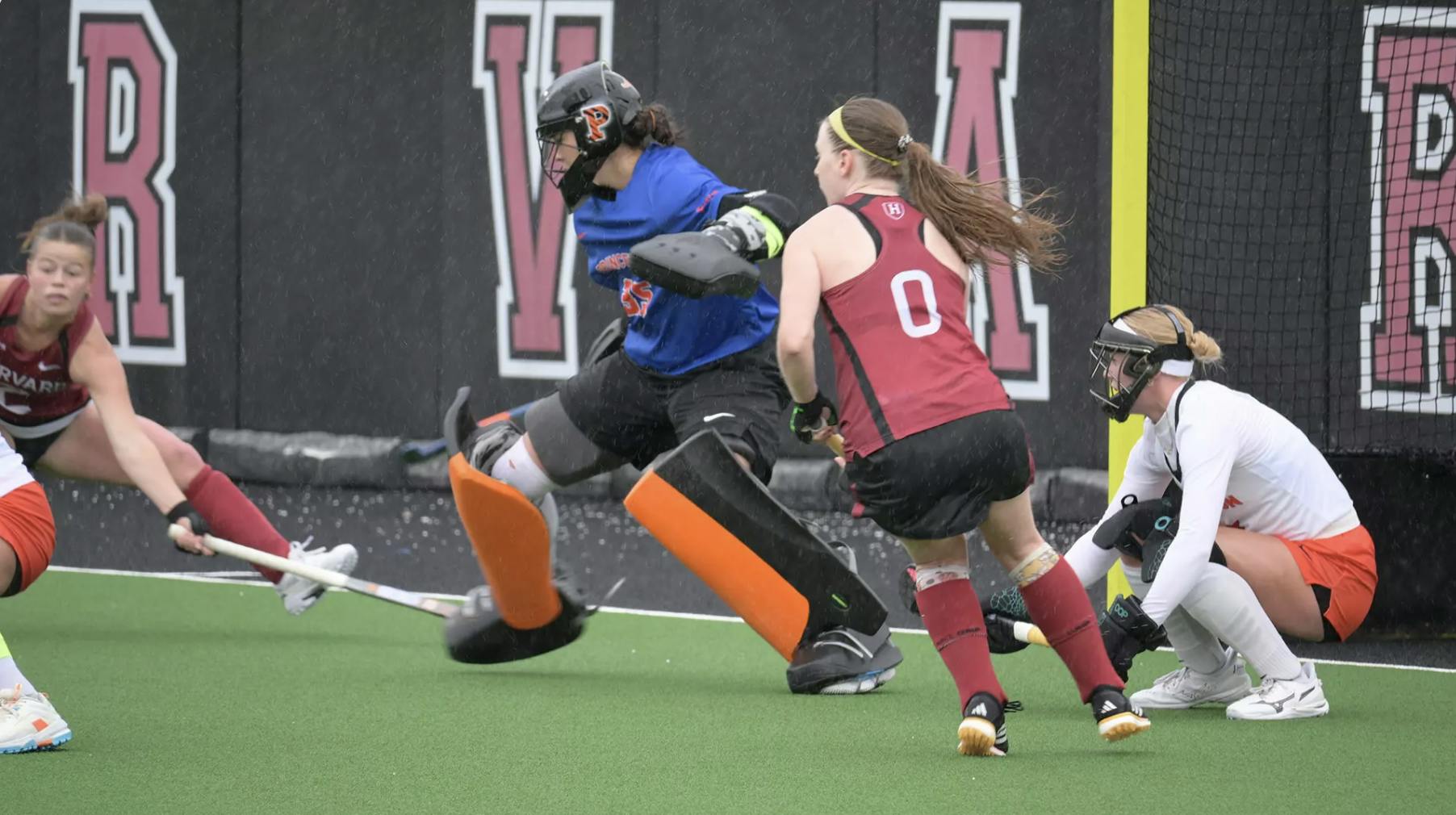2 players in red and black, 1 player in blue and black, and 1 player in white and orange play in front of the net.