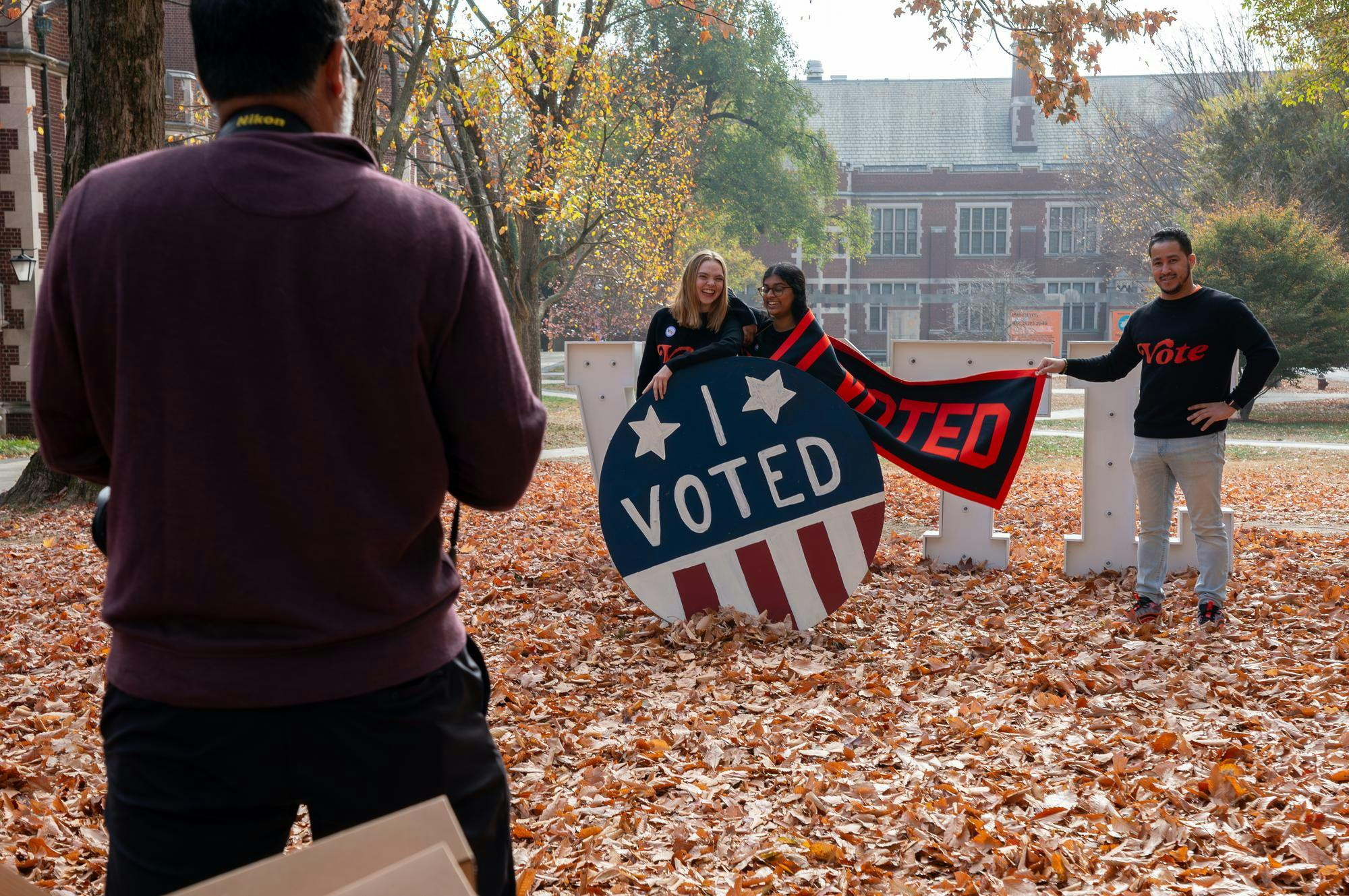 2 students and an administrator pose for photos in front of a large “I Voted” sign. Their photographer is in frame.