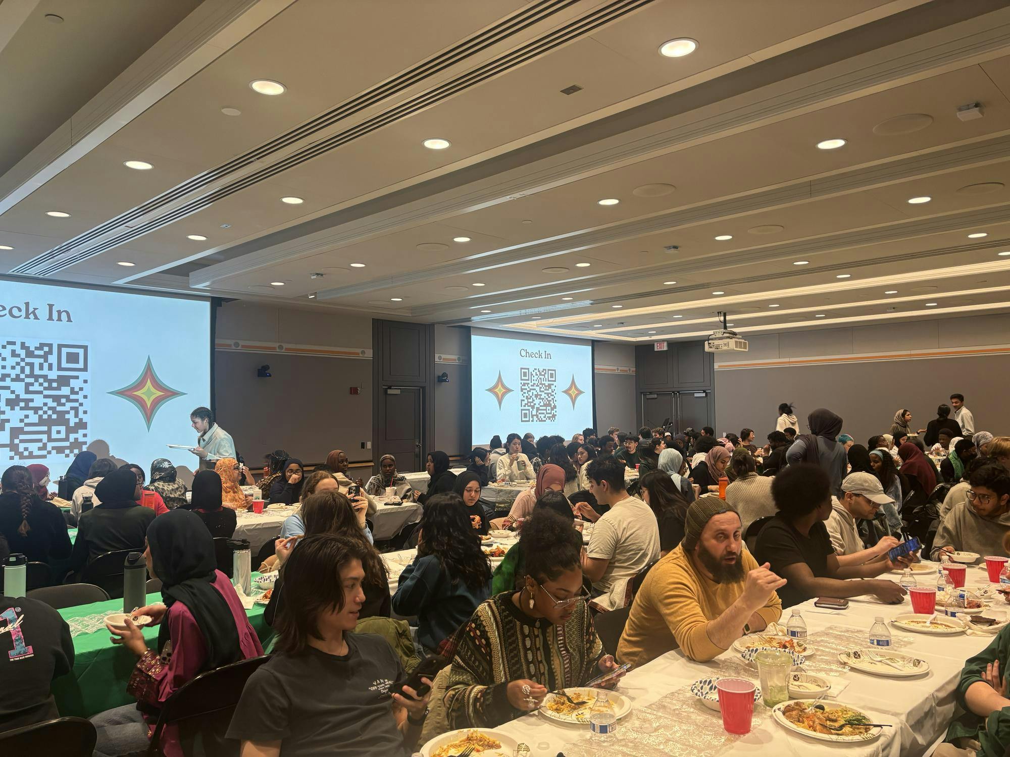 People sit in rows in a large room, eating from paper plates and talking. Projector screens at the back of the room display large QR codes for attendees to check-in.