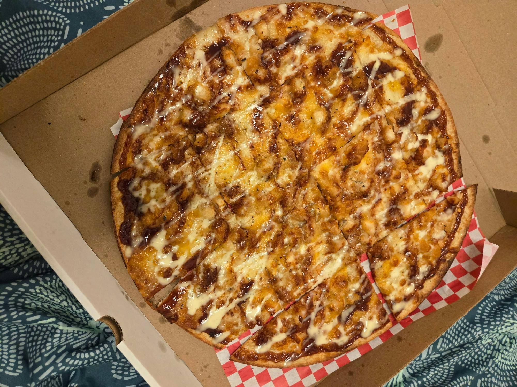 A pizza sits on top of red and white checkered paper. The pizza is inside of a cardboard box.