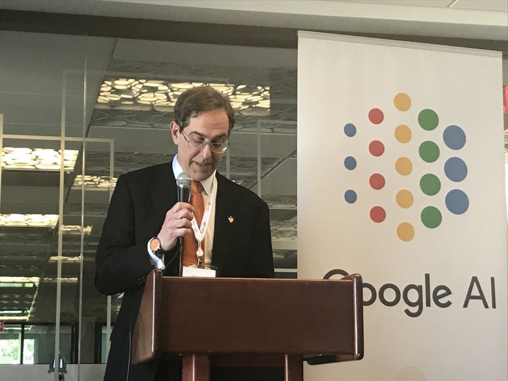 Princeton University President Christopher Eisgruber '83 stands in a suit and an orange tie behind a brown podium with a microphone in hand. A banner that says "Google AI" in blue, red, yellow, and green is next to him.