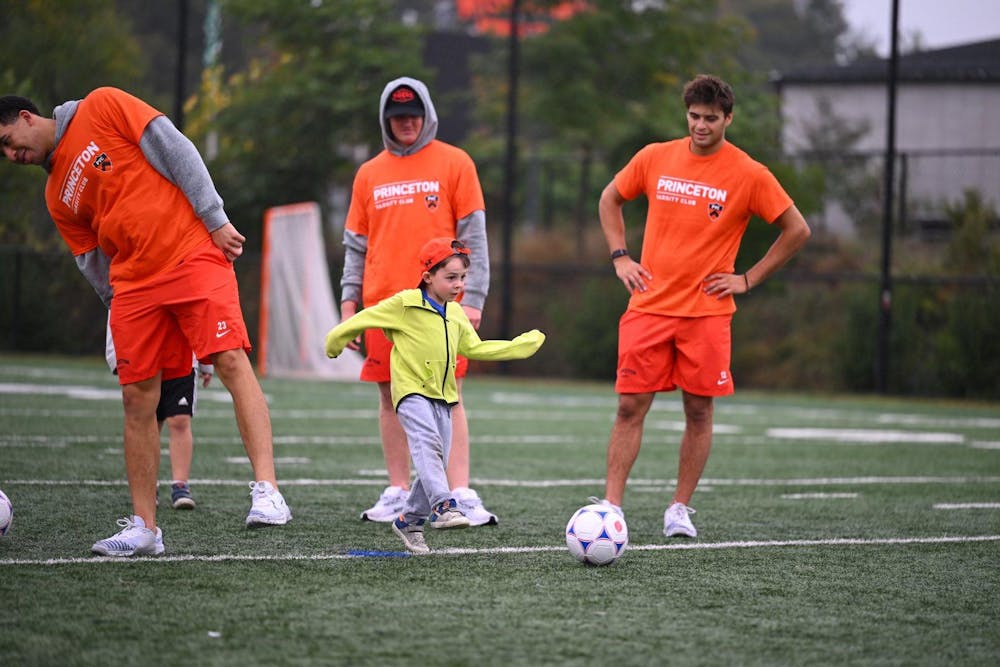 A young kid wearing a neon green jacket with gray sweatpants rocking an orange cap backward kicks a soccer ball as he is watched on by two young adults wearing primarily orange. 