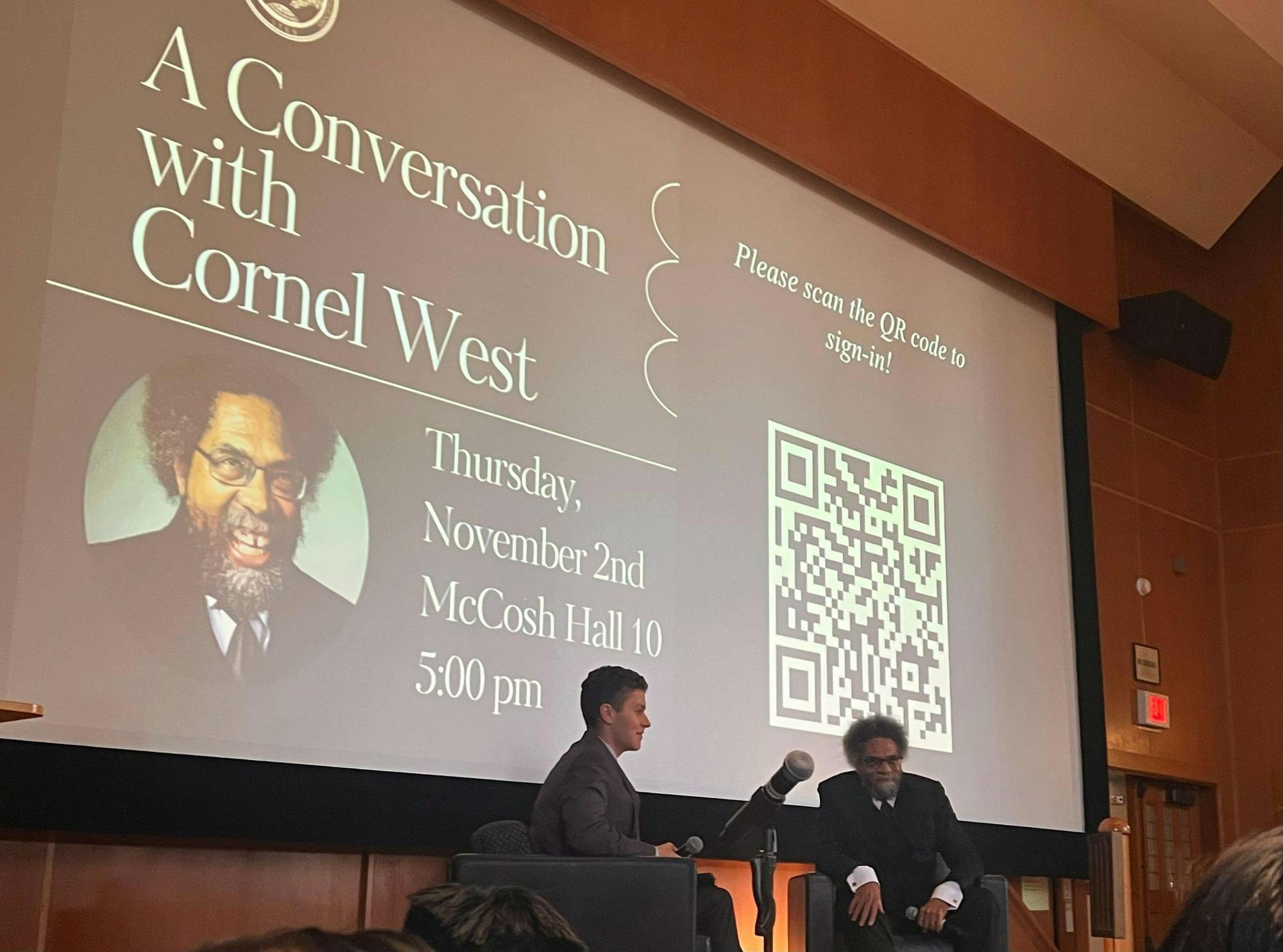 Two people sit in chairs on stage in front of screen projecting event information.