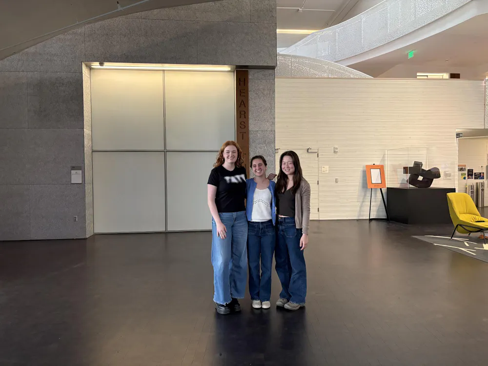 Three femme-appearing people wearing multicolored outfits are standing on a black hardwood floor.
