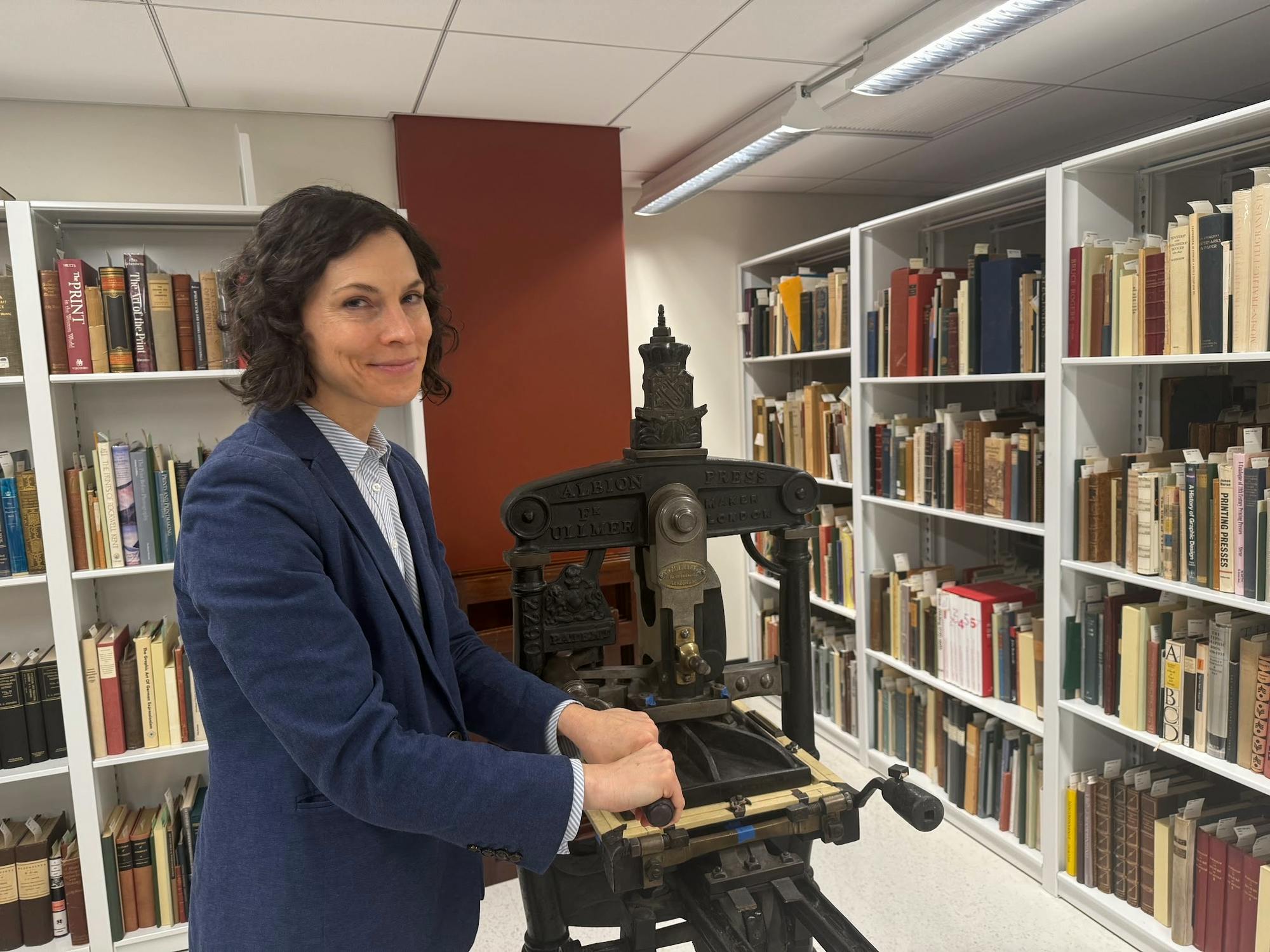 A woman in a blue blazer flanked by bookshelves stands next to an old, dark metal printing press, her hands on the lever.