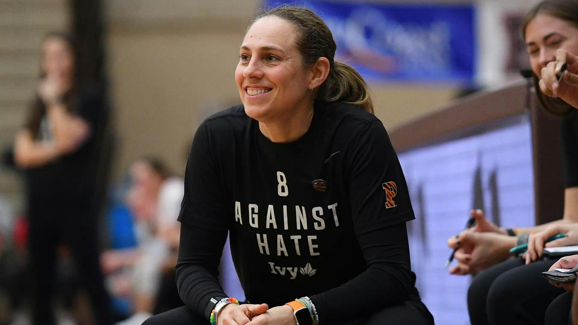 Woman in black shirt smiles on sideline of basketball game.
