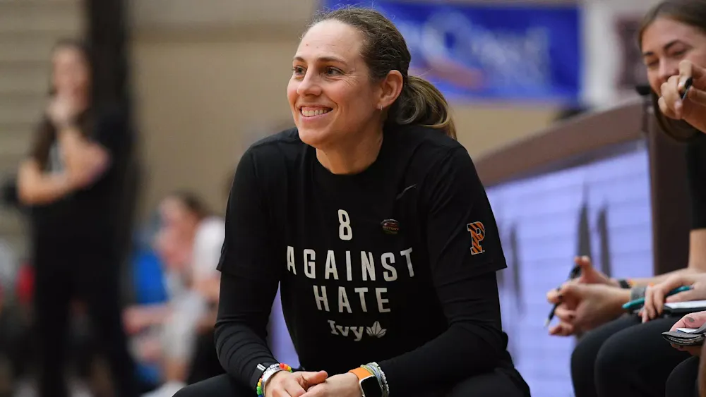 Woman in black shirt smiles on sideline of basketball game.