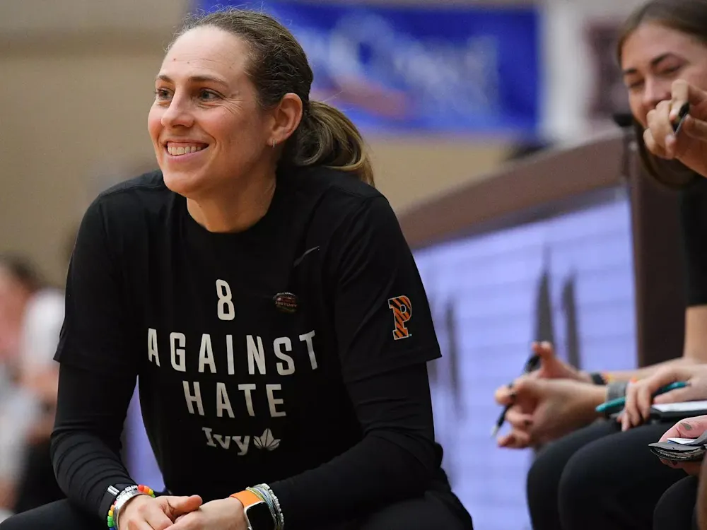 Woman in black shirt smiles on sideline of basketball game.