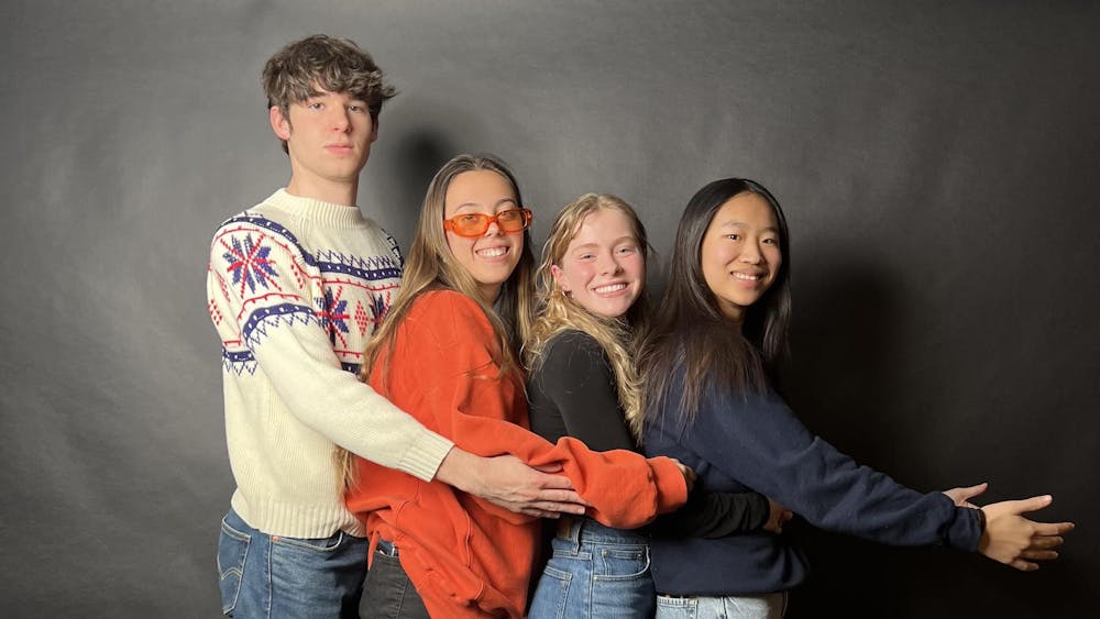 One male and three female students stand against a black background and face the camera while grabbing onto each others waists in a prom-like pose.