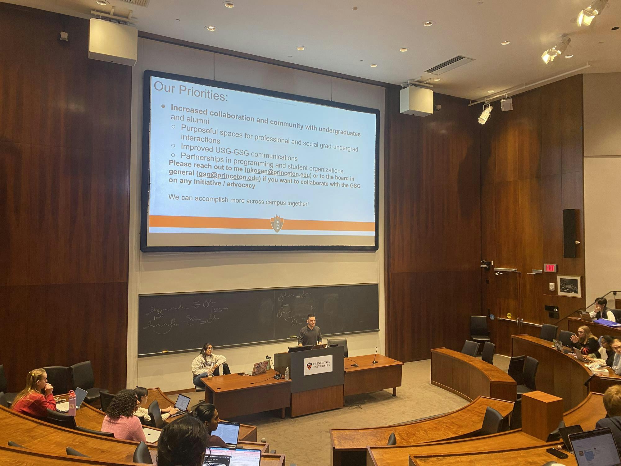 A large wood-paneled auditorium with a large projector screen titled “Our priorities,” with a bolded bullet point about increasing collaboration and community with undergraduates.