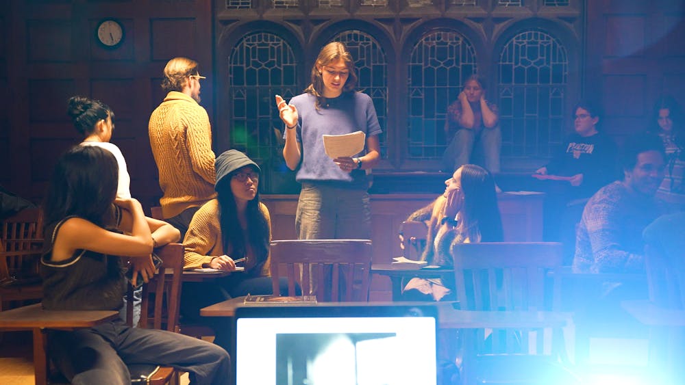 Person stands at the center of a classroom, reading a script, as cast members watch her. 