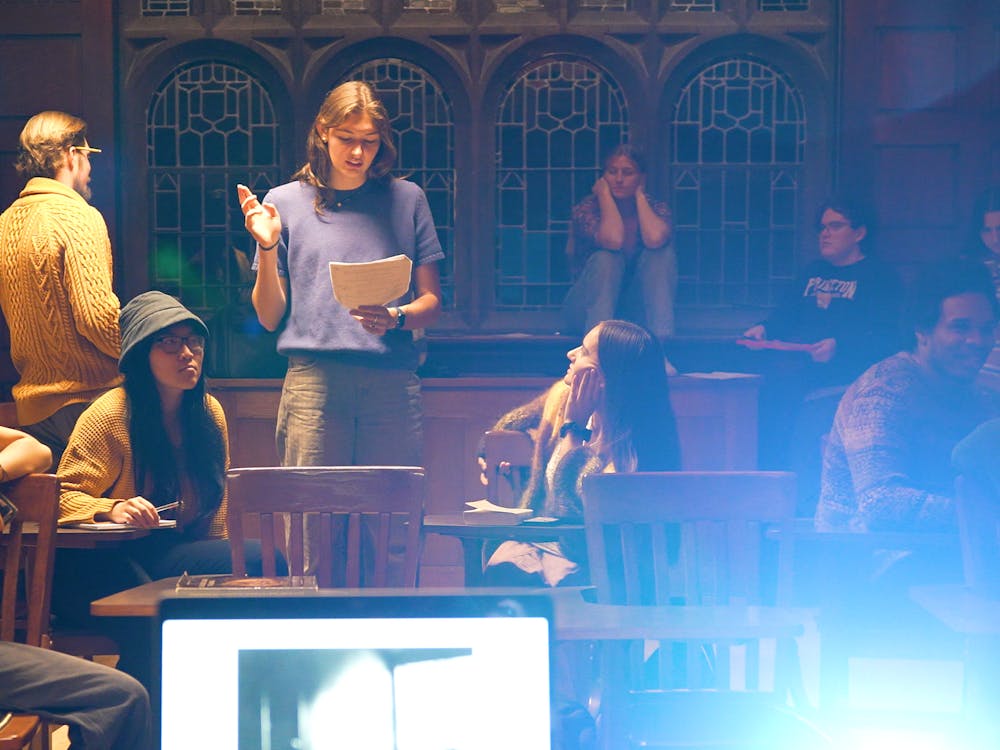 Person stands at the center of a classroom, reading a script, as cast members watch her. 