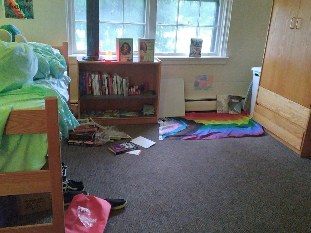 A single dorm room with a bed with green sheets on the left side and various items strewn on the floor, such as books, shoes, and the LGBTQ+ flag.