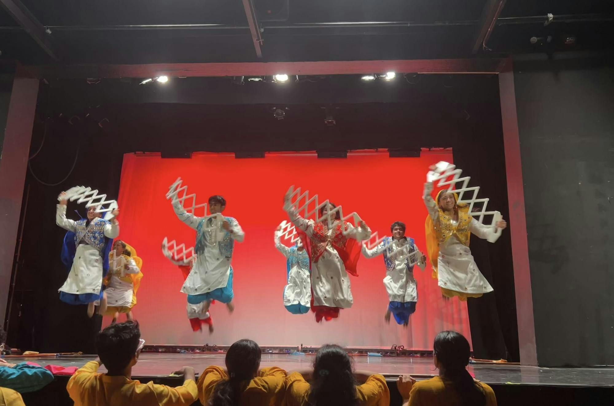 Dancers in multicolored outfits jump while holding a white prop against a red background.