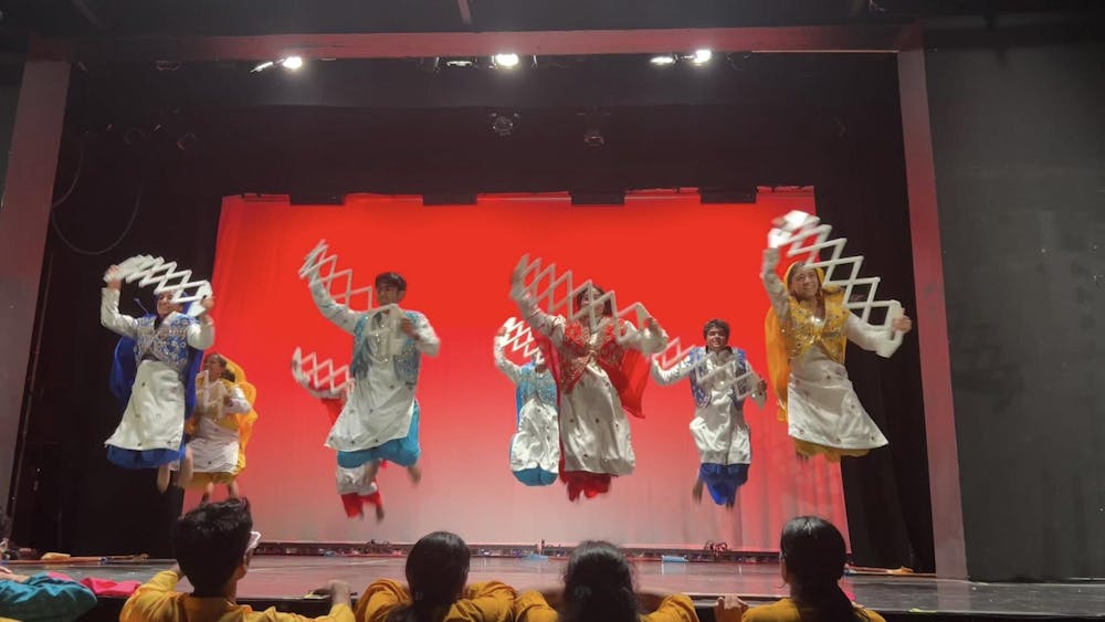 Dancers in multicolored outfits jump while holding a white prop against a red background.