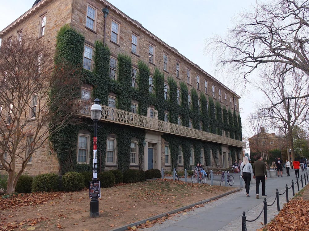 A brown brick building with ivy growing on it. A number of people are walking past the building.